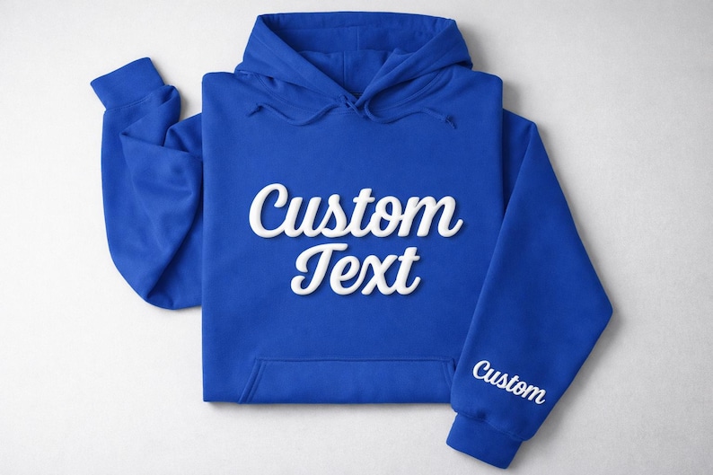 May include: Royal blue hoodie with the words "Custom Text" in white script across the front. The sleeve also has the word "Custom" in white script. The hoodie is laid flat on a white surface.