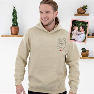May include: A beige hoodie with a front pocket and a hood. The hoodie features a black line drawing of a person holding a child, with the words "Daddy's Sunshine" and a red heart. A framed photo is on the right.