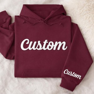 May include: A maroon hoodie with the word "Custom" in white script across the chest and on the left sleeve. The hoodie has a hood with drawstrings and a front pocket. The garment is folded on a white, fluffy surface.