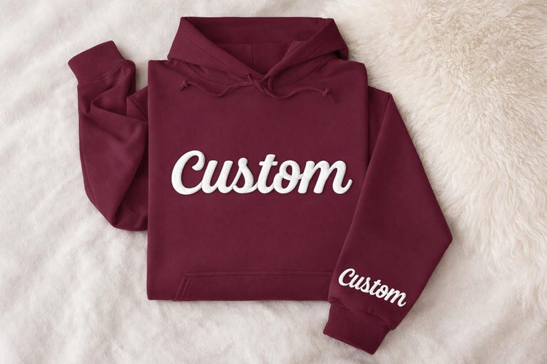 May include: A maroon hoodie with the word "Custom" in white script across the chest and on the left sleeve. The hoodie has a hood with drawstrings and a front pocket. The garment is folded on a white, fluffy surface.