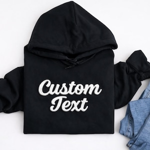 May include: Black hooded sweatshirt with the text "Custom Text" in a white, cursive font. The sweatshirt is paired with a pair of blue jeans. The hoodie has a drawstring and a front pocket.