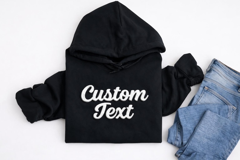 May include: Black hooded sweatshirt with the text "Custom Text" in a white, cursive font. The sweatshirt is paired with a pair of blue jeans. The hoodie has a drawstring and a front pocket.