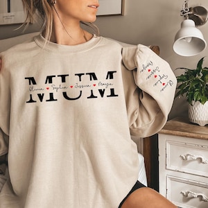 May include: Beige sweatshirt with the word "MUM" in black letters and the names "Olivia", "Sophia", "Jessica", and "Maya" written in cursive with a red heart between each name.