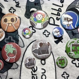 Pode incluir: Uma coleção de botões redondos e coloridos com vários desenhos de personagens de desenhos animados. Os botões apresentam uma variedade de cores, incluindo marrom, vermelho, verde, azul e cinza. Alguns botões têm um personagem com um design floral, outros um robô.