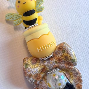 May include: A decorative pen featuring a yellow bee figure, a yellow honey pot with the word HUNNY printed on it, a floral patterned fabric bow in shades of orange and yellow, a clear bead with white flower accents, and a black fuzzy pom-pom.