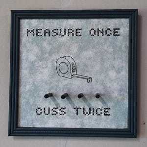 May include: A framed sign with the text "MEASURE ONCE CUSS TWICE". The sign features a tape measure illustration and four small pegs below the text. The frame is dark blue.