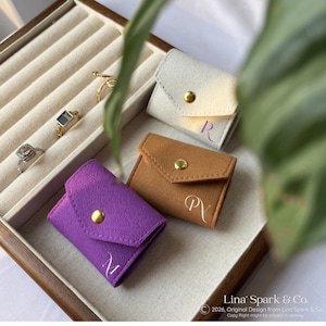 May include: A display of small, colorful ring pouches in purple, tan, and off-white. Each pouch has a gold snap closure and a single initial. Rings are visible in a jewelry tray. The text "Lina' Spark & Co." is at the bottom.