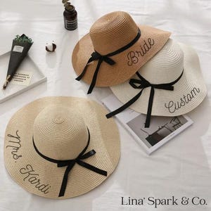 May include: Three straw sun hats with black ribbon accents. One hat is tan with "Bride" embroidered on the brim, another is white with "Custom", and the third is beige with "Mrs. Kardi". The hats are for a bridal party.