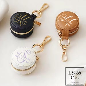 May include: Three personalized round zippered cases in black, tan, and white, each with a gold zipper and clasp. The cases feature custom initials and names in elegant script. The text "KAILY", "NANCY", and "SANDRA" are visible.