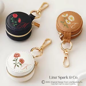 May include: Round, zippered keychains in black, brown, and white, each adorned with floral designs and a gold zipper. The black keychain features red roses and the letter "M". The brown keychain has daisies and the letter "E". The white keychain displays pink roses and the letter "K".