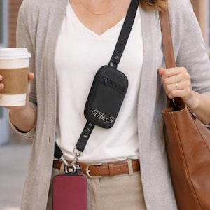 May include: A black crossbody phone case with a zipper closure and the cursive text "Mrs. S". The case is attached to a black strap and a burgundy phone case. The person is wearing a light gray cardigan and a white shirt.