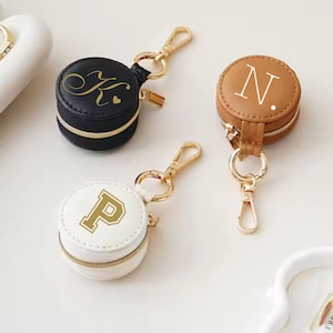 May include: Three round zippered pouches with gold keychains. The pouches are black, white, and brown. The black pouch has a gold "K" with a heart, the white pouch has a gold "P", and the brown pouch has a white "N".