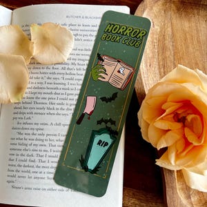 Könnte beinhalten: Ein grüner Lesezeichen mit dem Text "HORROR BOOK CLUB" und gruseligen Illustrationen eines Buches, eines Beils, Fledermäusen und einem Grabstein. Das Lesezeichen liegt auf einem offenen Buch mit einer Rose in der Nähe.