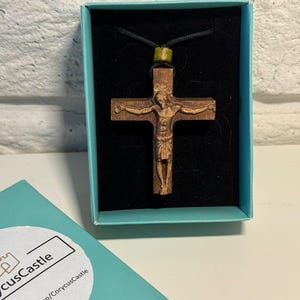 Pode incluir: Um crucifixo de madeira castanha com a figura esculpida de Jesus, suspenso num cordão preto com uma pequena conta de cor azeitona. O pendente é apresentado dentro de uma caixa azul-clara com um encaixe de veludo preto e um autocolante.