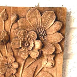 Op de afbeelding: Close-up van een houten snijwerk met gedetailleerde bloemmotieven. Het snijwerk toont grote, open bloemen met ingewikkelde bloemblaadjes, knoppen en bladeren. Het hout heeft een warme, natuurlijke tint, die de textuur en diepte benadrukt.