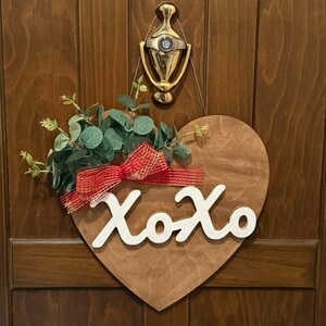 Op de afbeelding: Een hartvormig houten deurbord met de witte tekst XOXO, versierd met een rood en goudkleurig gaaslint en een bosje groene eucalyptusbladeren, hangend aan een goudkleurige deurklopper op een bruine houten deur.