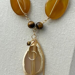 K&ouml;nnte beinhalten: Eine handgefertigte Halskette mit gro&szlig;en, bernsteinfarbenen Steinen, die mit Golddraht umwickelt sind. Die Halskette enth&auml;lt kleinere, runde, braune Perlen und einen Anh&auml;nger in Muschelform, der mit Goldketten und einem Goldring verziert ist. Der Schmuck wird auf neutralem Hintergrund pr&auml;sentiert.