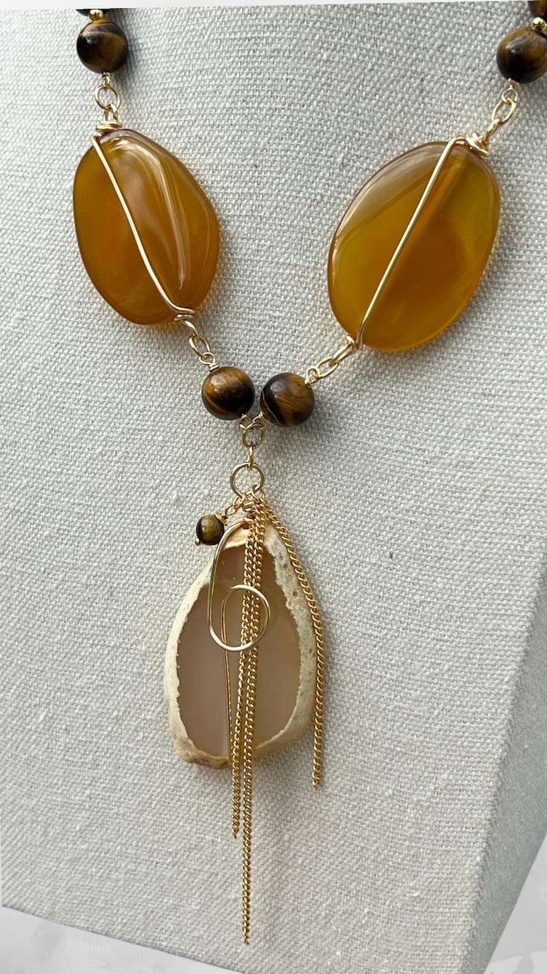 K&ouml;nnte beinhalten: Eine handgefertigte Halskette mit gro&szlig;en, bernsteinfarbenen Steinen, die mit Golddraht umwickelt sind. Die Halskette enth&auml;lt kleinere, runde, braune Perlen und einen Anh&auml;nger in Muschelform, der mit Goldketten und einem Goldring verziert ist. Der Schmuck wird auf neutralem Hintergrund pr&auml;sentiert.