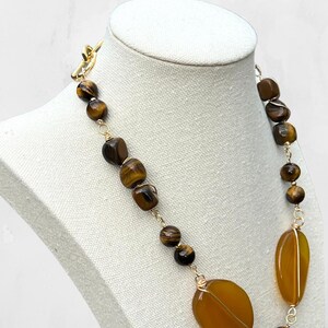 K&ouml;nnte beinhalten: Goldfarbene Halskette mit braunen Tigerauge-Perlen und gro&szlig;en, ovalen, bernsteinfarbenen Steinen. Die Halskette wird auf einer wei&szlig;en Mannequin-B&uuml;ste pr&auml;sentiert, um ihr Design und ihre Farbvariationen hervorzuheben. Mit einem Knebelverschluss.