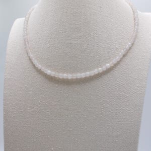May include: A delicate necklace composed of small, pale pink beads. The necklace is displayed on a white mannequin, showcasing its simple elegance. The beads appear to be uniform in size, creating a smooth, continuous strand. This piece is suitable for everyday wear.