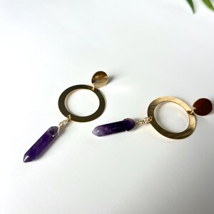 May include: A pair of gold hoop earrings with a small gold disc at the top and a purple amethyst crystal dangling from the bottom.