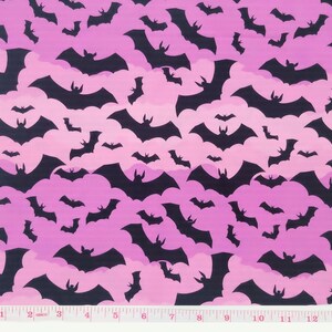 May include: Pink fabric with a repeating pattern of black bats. The fabric is a gradient of pink shades, with a darker pink at the top and a lighter pink at the bottom. The bats are all facing the same direction, with their wings spread out. The fabric is measured in inches.