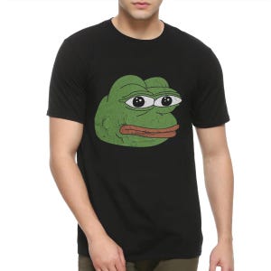 Op de afbeelding: Zwart T-shirt met een verweerde groene kikkergezichtsafbeelding. De kikker heeft grote ogen en een bruine mond. De afbeelding is gecentreerd op de voorkant van het shirt. Het T-shirt is gemaakt van een zacht materiaal.