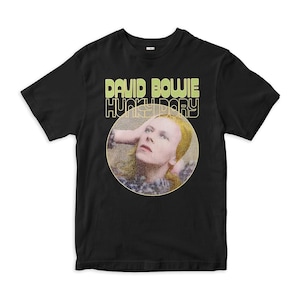 Könnte beinhalten: Schwarzes T-Shirt mit einem Grafikdruck von David Bowie aus dem Album "Hunky Dory". Die Grafik ist in einem Kreis mit gelbem Hintergrund und zeigt Bowie mit seinen Haaren, die seine Augen bedecken.