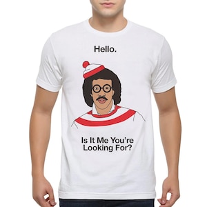 Puede incluir: Camiseta blanca con una ilustración de dibujos animados de un hombre con un suéter a rayas rojas y blancas, un gorro rojo y blanco y gafas. El texto "Hello." está por encima de la ilustración y el texto "Is It Me You're Looking For?" está por debajo de la ilustración.
