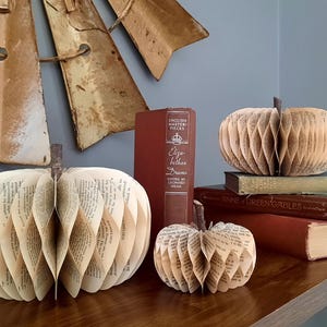 May include: Three paper pumpkins made from book pages sit on a wooden table with a stack of books. The pumpkins have brown stems and are in various sizes. A weathered windmill blade hangs in the background.