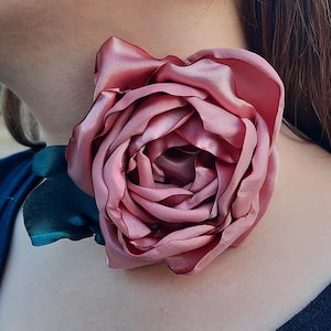 May include: A large, pink satin rose flower, with a teal green fabric leaf, is worn as a choker necklace.