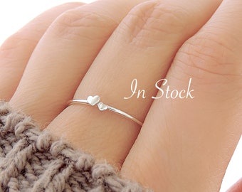 Può includere: Un delicato anello d'argento con due piccoli accenti a forma di cuore. L'anello è indossato su un dito.