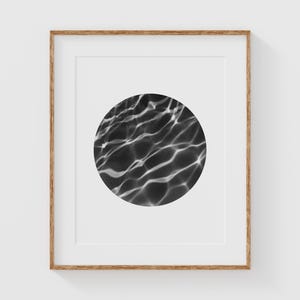 Peut inclure: Impression d'art encadrée avec un design abstrait en noir et blanc. L'œuvre présente une image circulaire d'eau ondulante, créant une sensation de profondeur et de mouvement. L'impression est posée sur un fond blanc et encadrée dans un ton bois naturel.
