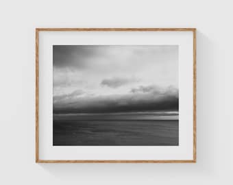 May include: A black and white photograph depicting a stormy sky over a calm body of water. The clouds are dark and dramatic, while the water is smooth and reflective.