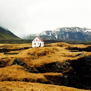 Red roofed white house on the edge of a cliff, with snow-capped mountains in the background. This photography print was shot on the west coast of Iceland