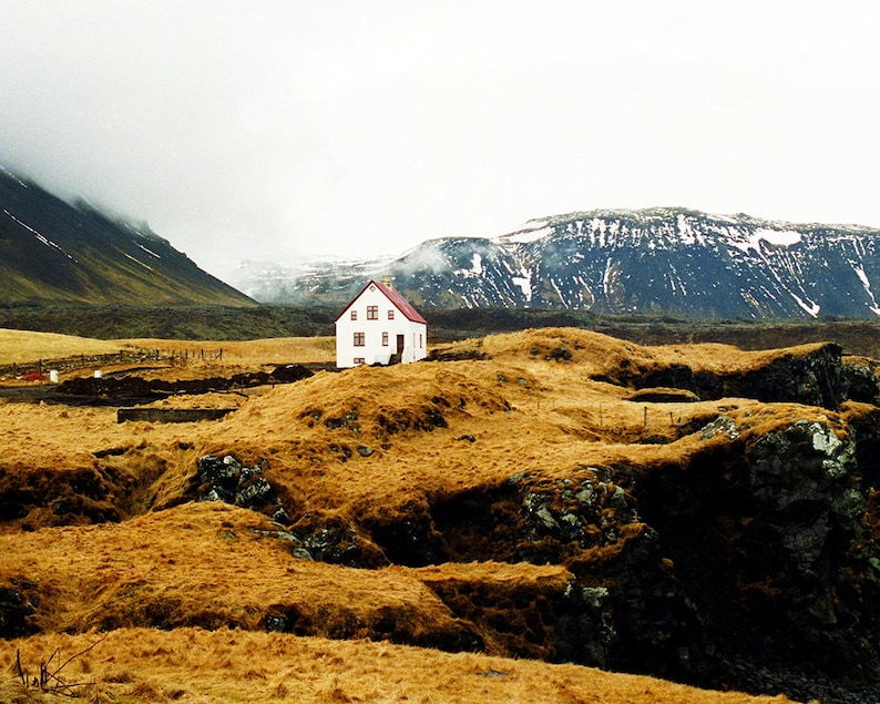 Red roofed white house on the edge of a cliff, with snow-capped mountains in the background. This photography print was shot on the west coast of Iceland