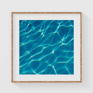 May include: A framed print featuring an abstract image of rippling water. The water is a deep, vibrant blue with highlights of white and lighter blue, creating a sense of depth and movement. The artwork is framed in a light brown wooden frame.
