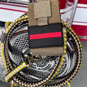 Könnte beinhalten: Ein braunes und beigefarbenes Paracord-Lanyard mit einem schwarzen und roten Patch, auf dem "Thin Red Line" steht. Das Lanyard ist an einer braunen Tasche mit einem schwarzen Clip befestigt.