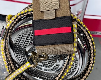 May include: A tan tactical radio pouch featuring a black rectangular patch with a horizontal red stripe. The pouch is attached to a braided paracord lanyard in black, tan, white, and yellow, finished with a brass-toned metal swivel clip.