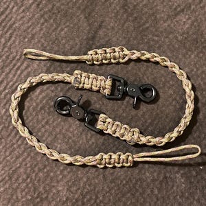 May include: A tan and brown camouflage patterned paracord lanyard featuring a braided design with two black metal swivel lobster claw clasps. The cord is looped at both ends with reinforced knot sections, creating a continuous, flexible strap.