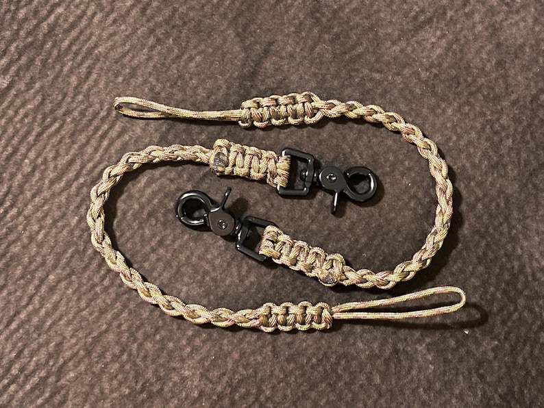 May include: A tan and brown camouflage patterned paracord lanyard featuring a braided design with two black metal swivel lobster claw clasps. The cord is looped at both ends with reinforced knot sections, creating a continuous, flexible strap.