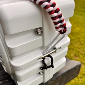 May include: A white, rigid, rectangular storage container featuring a thick, braided paracord handle in a red, white, and black pattern. The handle is attached to a silver metal D-ring secured by a small black metal shackle on the side of the unit.