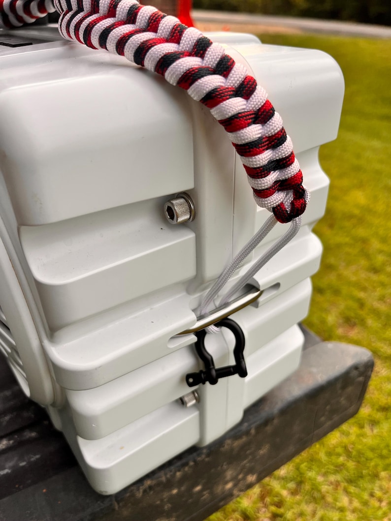 May include: A white, rigid, rectangular storage container featuring a thick, braided paracord handle in a red, white, and black pattern. The handle is attached to a silver metal D-ring secured by a small black metal shackle on the side of the unit.