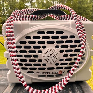 May include: A grey Turtlebox speaker with a black, red, and white braided strap. The speaker has a black handle and the text "TURTLEBOX" on the front. The speaker is on a dark surface.