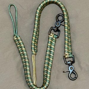 May include: A braided paracord lanyard featuring a camouflage pattern of olive green and tan colours. The cord is finished with a loop at one end and two dark metal swivel trigger snap hooks attached to the other end for secure fastening.