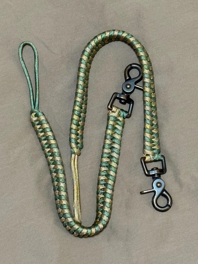 May include: A braided paracord lanyard featuring a camouflage pattern of olive green and tan colours. The cord is finished with a loop at one end and two dark metal swivel trigger snap hooks attached to the other end for secure fastening.