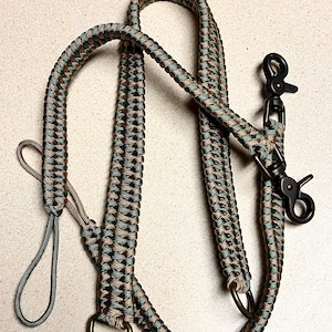 May include: A multi-purpose lead crafted from braided paracord in a camouflage pattern of dark green, brown, and tan. The lead features two dark metal swivel trigger snaps, two metal D-rings, and a small loop handle at one end for control.