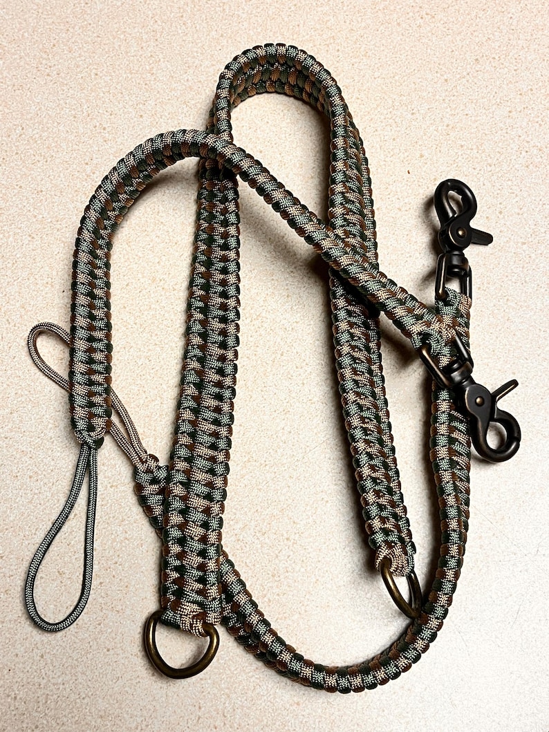 May include: A multi-purpose lead crafted from braided paracord in a camouflage pattern of dark green, brown, and tan. The lead features two dark metal swivel trigger snaps, two metal D-rings, and a small loop handle at one end for control.