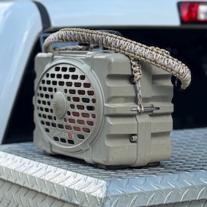 May include: A tan Turtlebox portable speaker featuring a rugged, rectangular design with a front-facing grille and a custom-woven, multi-coloured paracord handle attached to the top, showcasing a durable construction and a prominent brand logo.