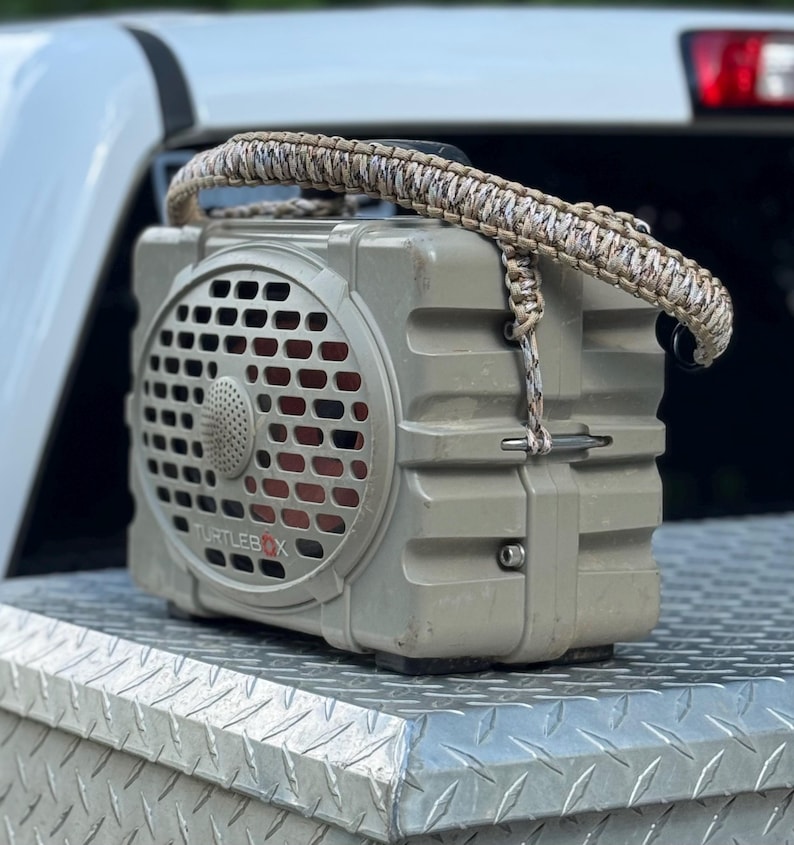 May include: A tan Turtlebox portable speaker featuring a rugged, rectangular design with a front-facing grille and a custom-woven, multi-coloured paracord handle attached to the top, showcasing a durable construction and a prominent brand logo.